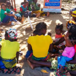 A group of women and children sit in a circle on the ground outdoors, some wearing colorful headscarves and clothing, engaged in a community discussion near a standing sign under the shade of trees.
