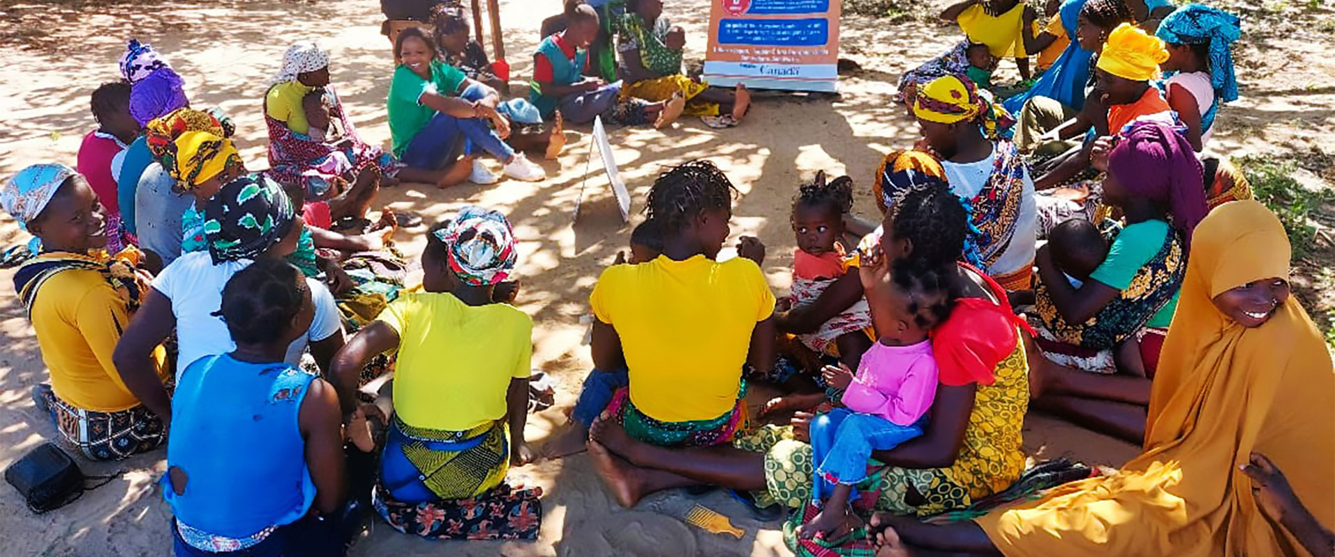 A group of women and children sit in a circle on the ground outdoors, some wearing colorful headscarves and clothing, engaged in a community discussion near a standing sign under the shade of trees.