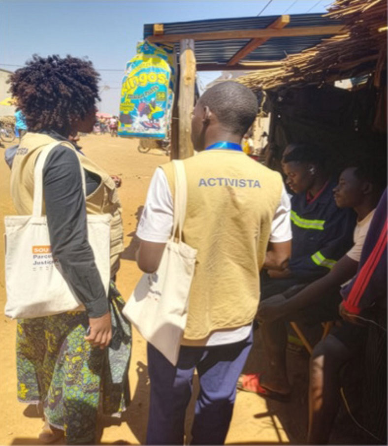 Two people wearing beige vests and holding tote bags, one labeled "ACTIVISTA," talk to two seated men in a shaded area outside in a sunny, rural setting with a “Lingos” sign in the background.