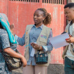 Two aid workers wearing beige vests speak with a woman who has a scarf over her head. One worker holds a clipboard while the other gestures, standing outside near a red gate and a stone wall.