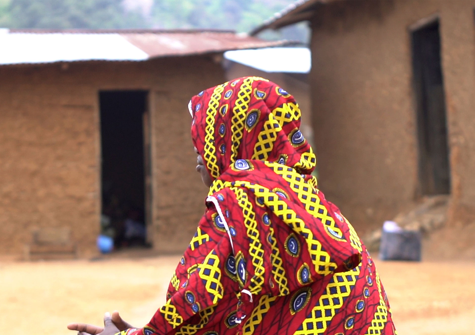 A person wearing a vibrant red, yellow, and black patterned garment sits outdoors, facing away from the camera, with earthen buildings in the background.