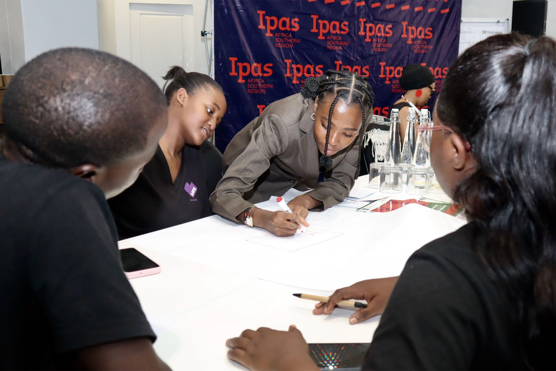 Four young adults sit around a table working together. One person leans over to write on a large sheet of paper. They are in a room with an "Ipas" banner in the background.