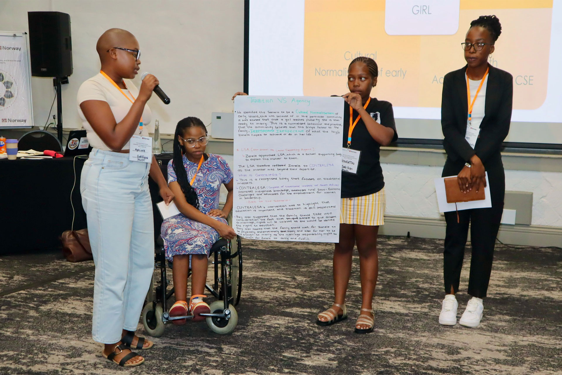 Four young people present at an event; one speaks with a microphone, one sits in a wheelchair, another holds a large paper with notes, and the fourth stands holding a tablet. A presentation screen is in the background.