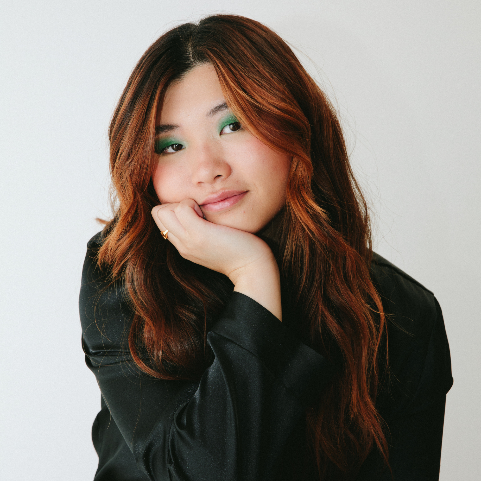 A young woman with long, wavy auburn hair and green eyeshadow rests her chin on her hand, wearing a black top and smiling gently against a plain white background.