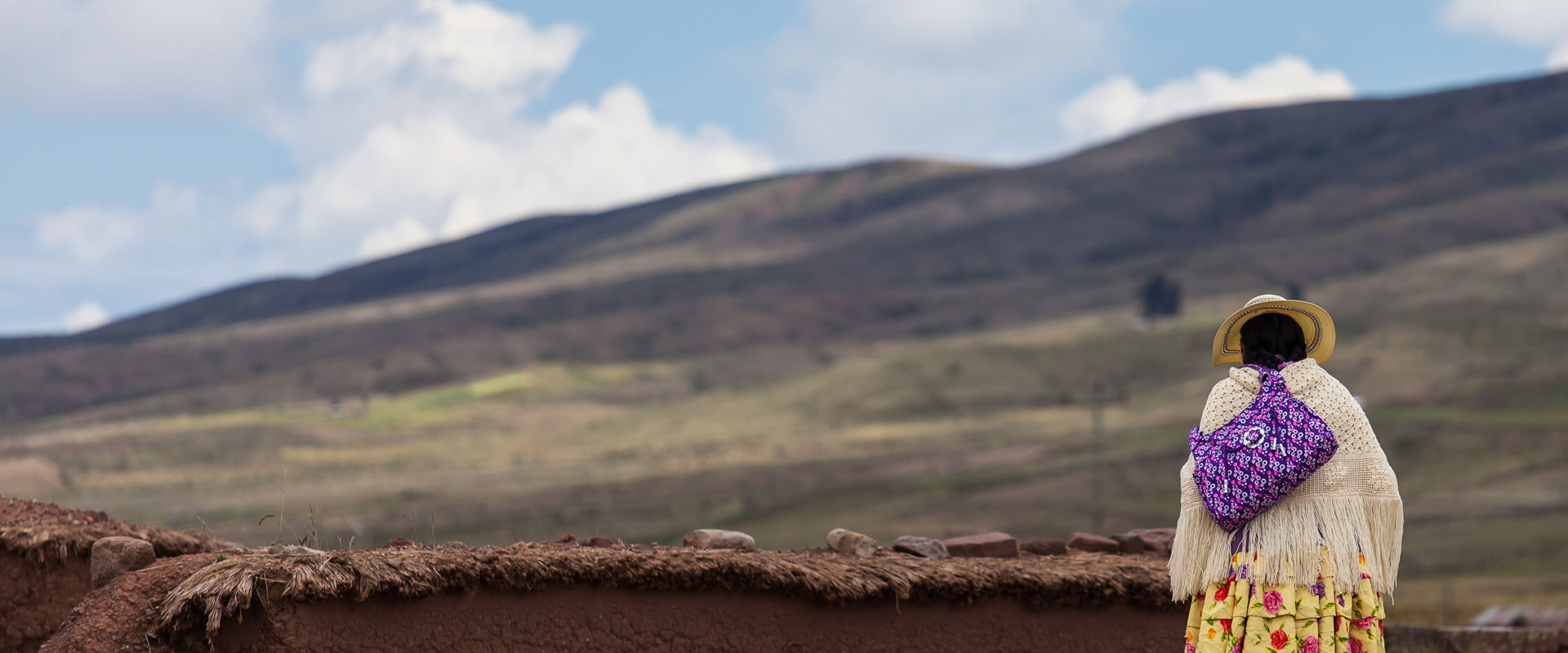 A woman in traditional clothing and a yellow hat stands by an adobe wall, facing distant mountains under a partly cloudy sky.