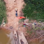 A person stands on a dirt path near a riverbank, possibly engaged in climate change research in Bolivia, with another person and scattered items nearby. The scene is surrounded by green vegetation and viewed from above.