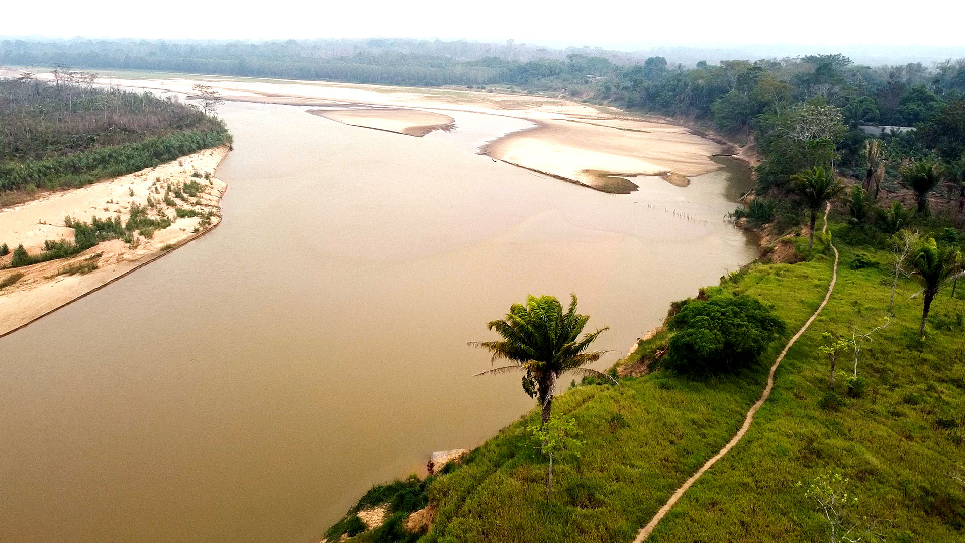 A winding river with sandy banks flows through a green landscape. There are trees and grass along the riverbank, with a narrow path visible beside the water under a cloudy sky.