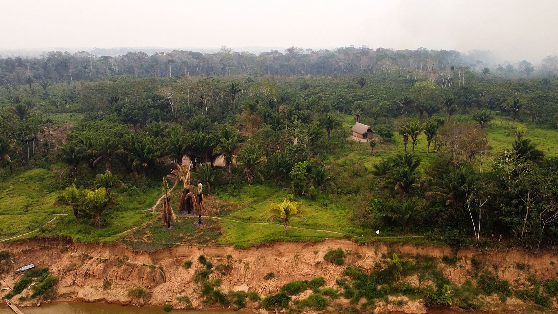 Aerial view of a dense green rainforest with scattered small huts and a riverbank with exposed soil and erosion in the foreground. The sky appears hazy.