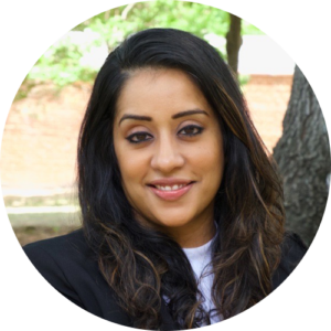 Headshot of Jina Dhillon, a woman with long dark hair and a black blazer smiles at the camera outdoors, with trees and greenery in the blurred background.