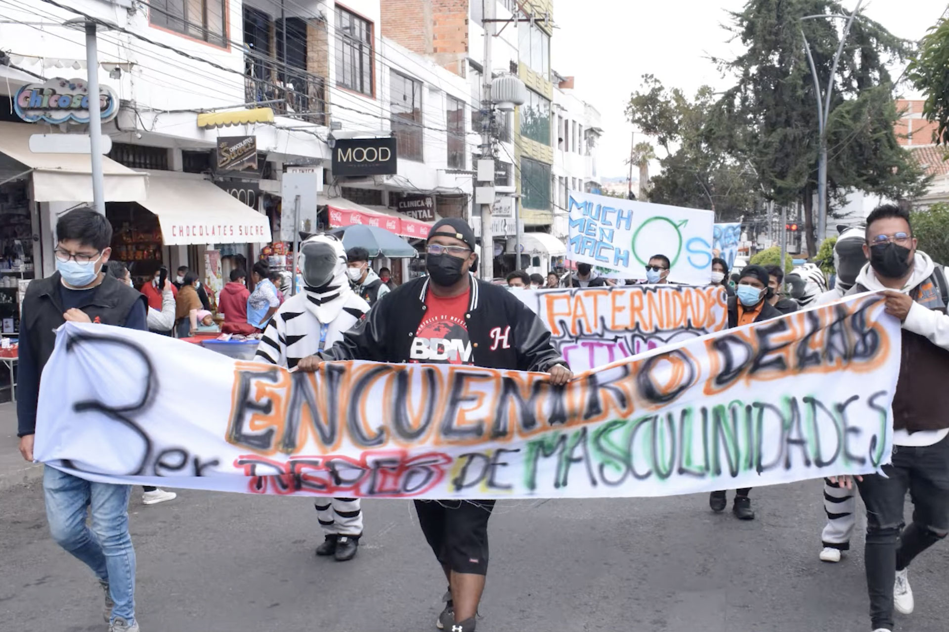 A group of people march outdoors, some in costumes, holding a colorful banner that reads “Encuentro de las Diversidad de Masculinidades.” Shops and bystanders are visible in the background.