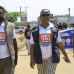 Four people walk on a street wearing “Stop Ebola” shirts; one holds a megaphone. Buildings, cars, and more people with a banner are visible in the background. The scene appears to be part of an awareness campaign.