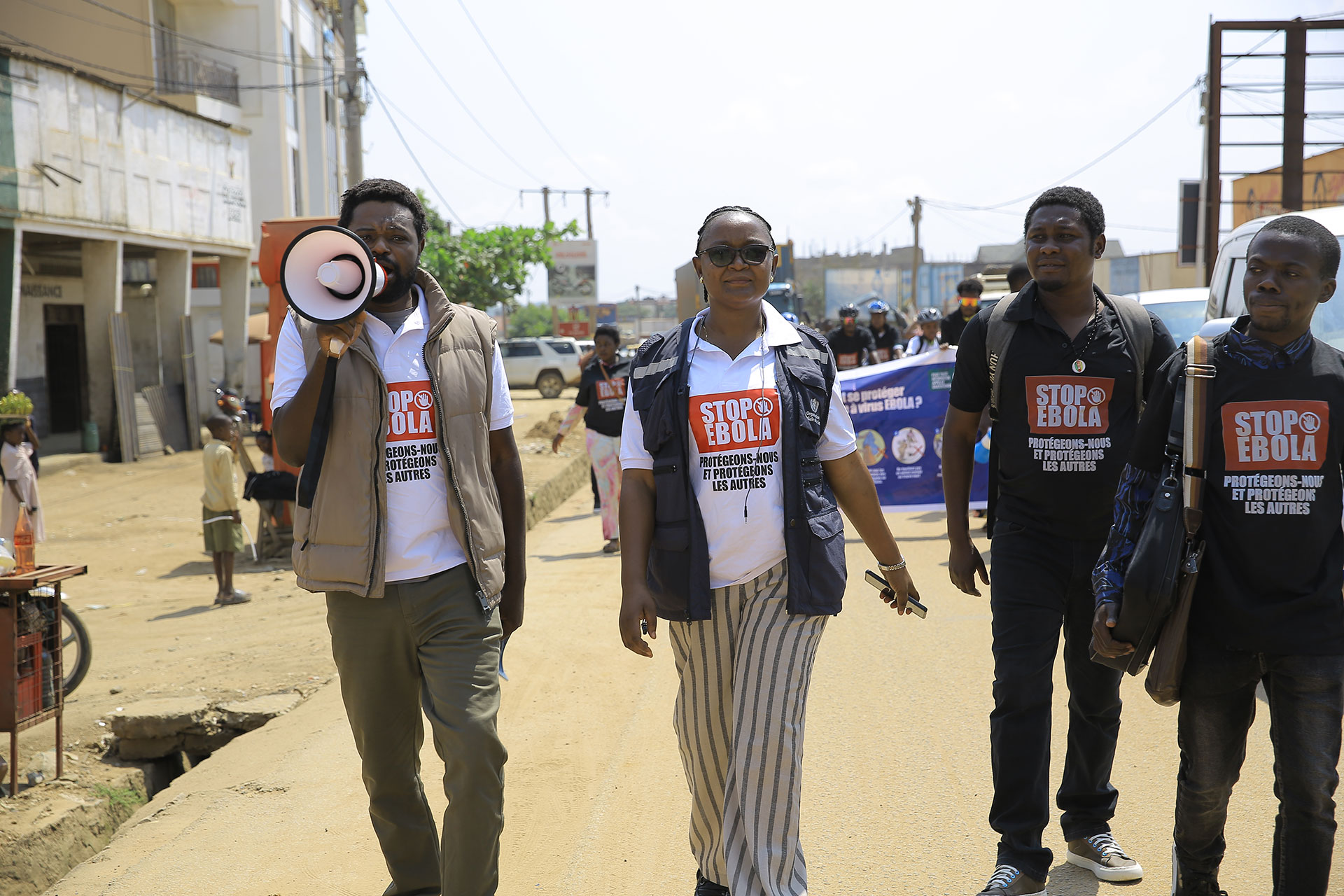 Four people walk down a street, with one speaking into a megaphone. They wear shirts that say “STOP EBOLA,” participating in a public awareness campaign. Buildings, a banner, and other individuals are visible in the background.