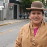 Portrait of former Bolivian Senator Virginia Velasco, a woman wearing a brown hat and patterned shawl smiles at the camera while standing on a city street, with people walking in the background.