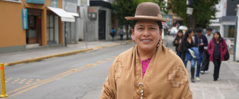 Portrait of former Bolivian Senator Virginia Velasco, a woman wearing a brown hat and patterned shawl smiles at the camera while standing on a city street, with people walking in the background.