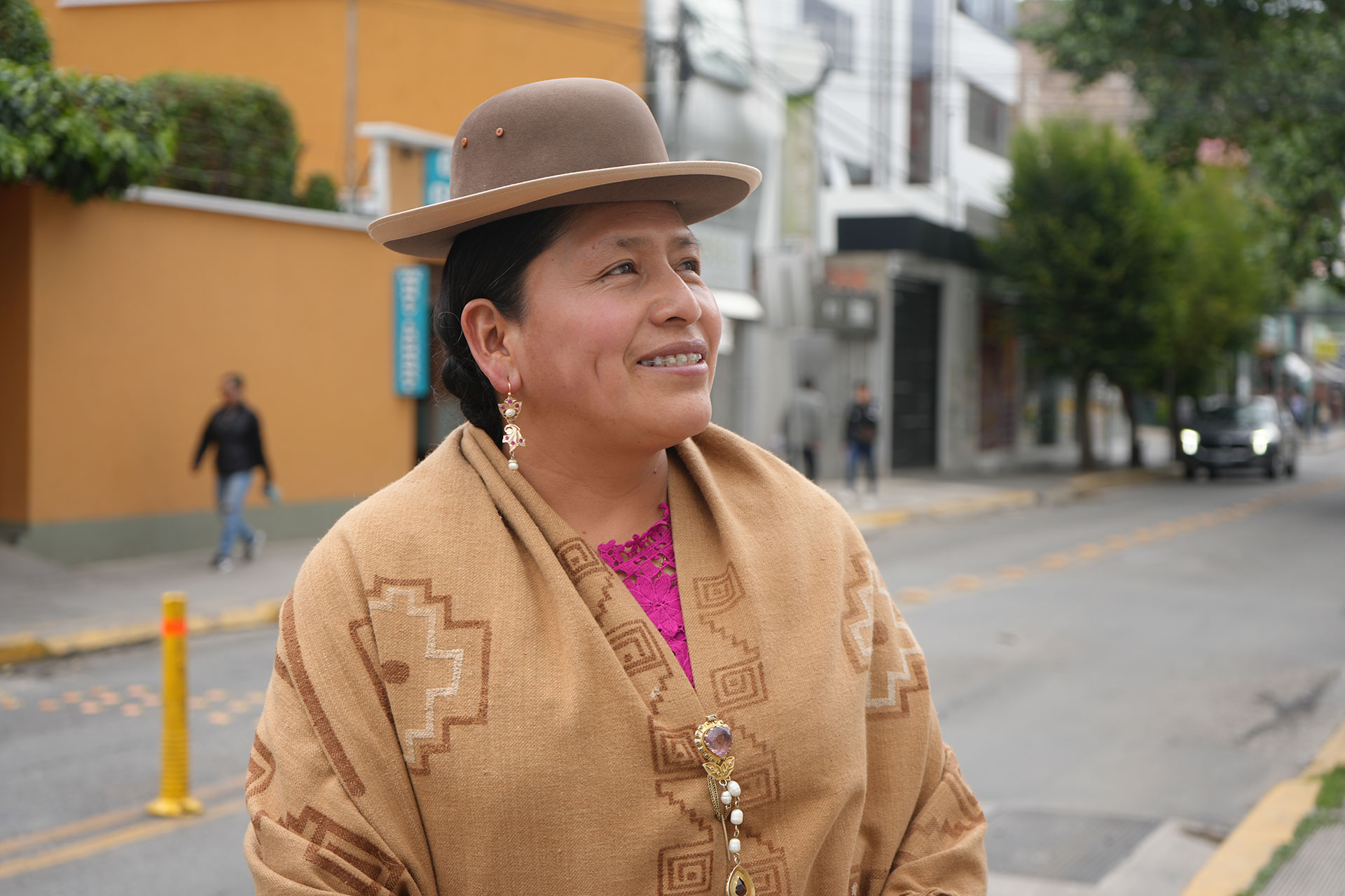 Portrait of former Bolivian Senator Virginia Velasco, a woman wearing a brown hat and patterned shawl smiles at the camera while standing on a city street, with people walking in the background.