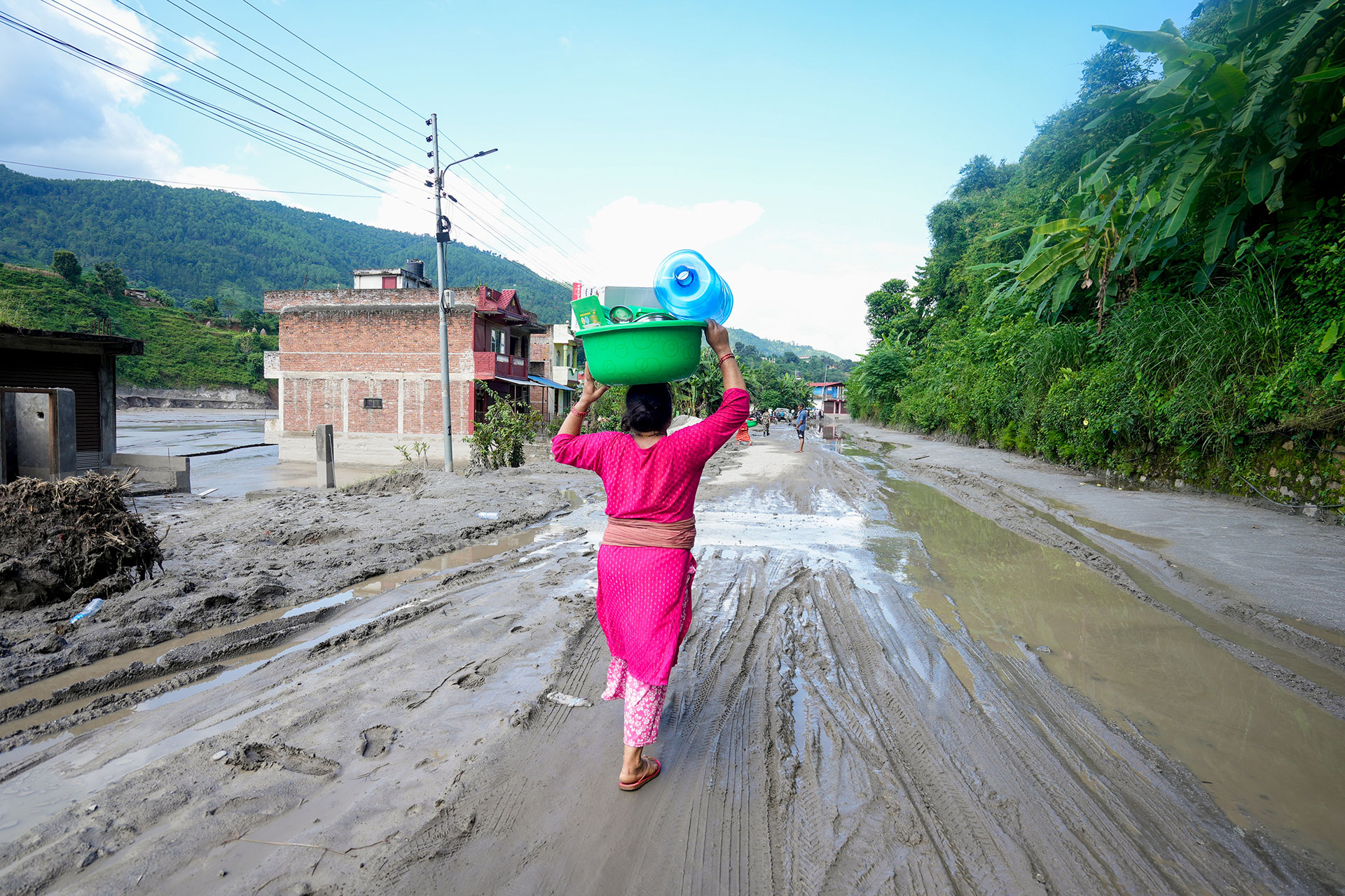 A woman in a pink outfit carries a green basket with items on her head, walking barefoot along a muddy, flooded road lined with trees and buildings in a rural area.