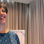 A woman with short dark hair smiling while speaking at a presentation with an Ipas banner in the background.