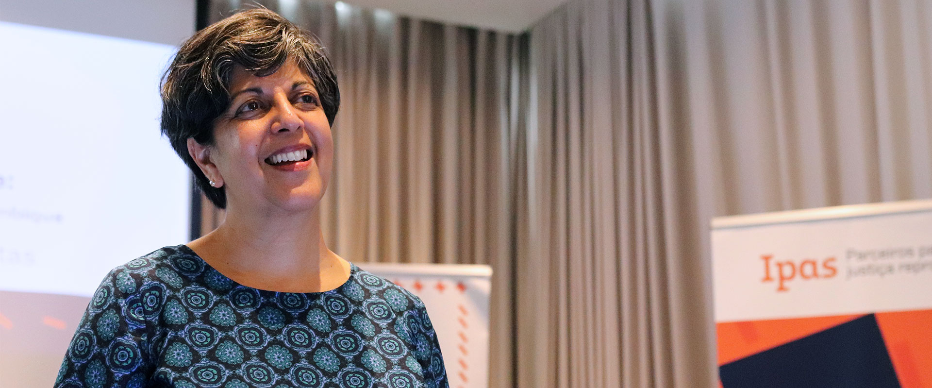 A woman with short dark hair smiling while speaking at a presentation with an Ipas banner in the background.
